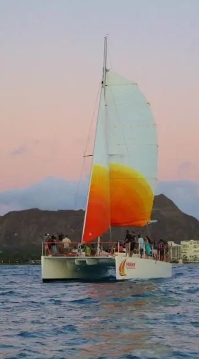 Sunset catamaran with a bright orange-yellow sail cruising off Waikiki with Diamond Head crater and Honolulu skyline in the background