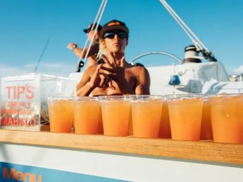 Row of orange tropical cocktails in plastic cups on a sailboat bar with a shirtless, sunglasses-wearing crew member, tip jar visible, and bright blue sky