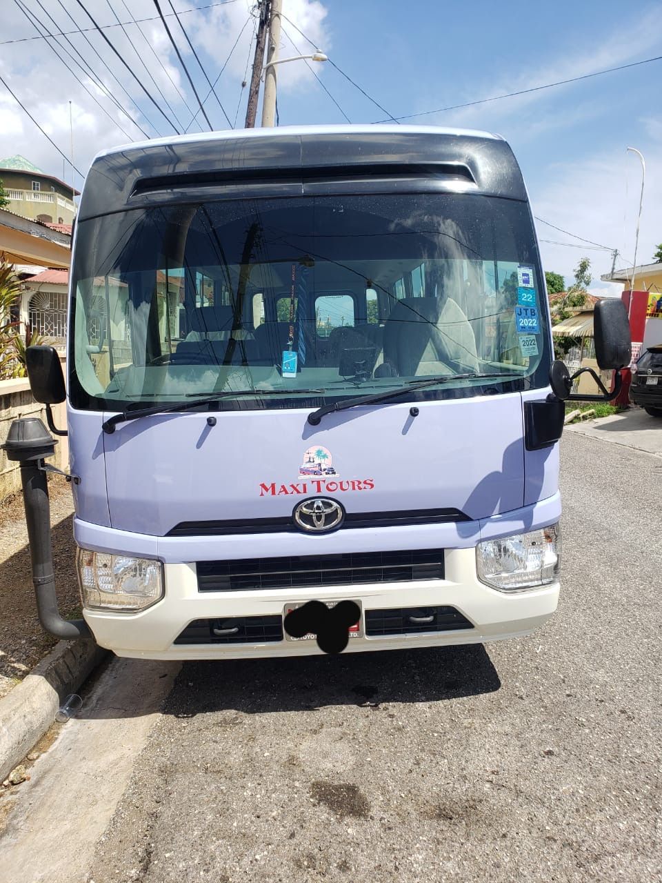 Front view of a light purple Toyota minibus tour shuttle parked on a sunny residential street, showing windshield inspection stickers, side mirrors, headlights, and overhead utility poles.