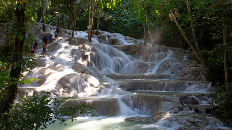 Multi-tiered limestone waterfall cascading into turquoise pools amid lush tropical forest, sunlight filtering through trees as visitors wade on the rocky steps