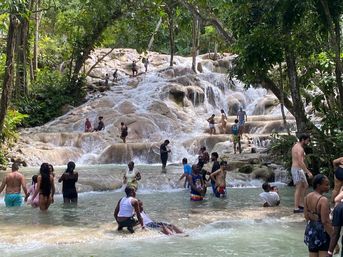 Crowded tropical waterfall with multi-tiered limestone cascades and turquoise pools, people wading, swimming and climbing the falls amid lush green rainforest