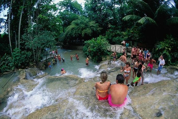 Couple perched on a rocky ledge overlooking a cascading tropical waterfall and natural pools, tourists swimming and exploring in a lush rainforest setting