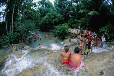 Couple perched on a rocky ledge overlooking a cascading tropical waterfall and natural pools, tourists swimming and exploring in a lush rainforest setting