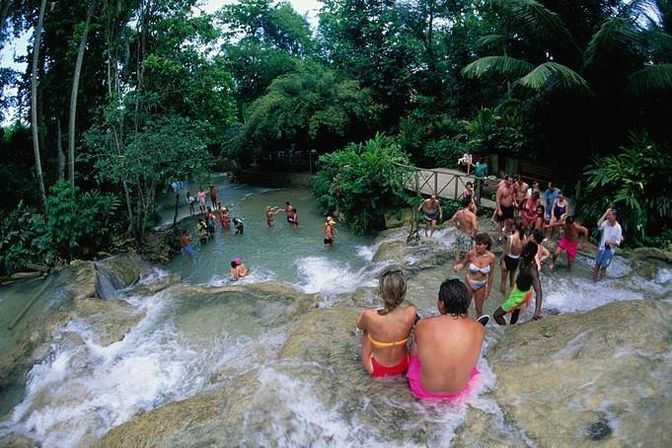 Couple perched on a rocky ledge overlooking a cascading tropical waterfall and natural pools, tourists swimming and exploring in a lush rainforest setting