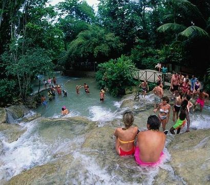 Couple perched on a rocky ledge overlooking a cascading tropical waterfall and natural pools, tourists swimming and exploring in a lush rainforest setting
