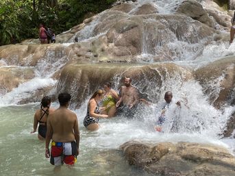 Group of people in swimsuits splashing and wading beneath a terraced tropical waterfall, standing on smooth limestone cascades and shallow natural pools