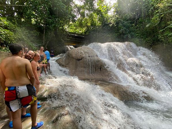 Group of people in swimsuits climbing a multi-tier tropical waterfall over smooth limestone cascades surrounded by lush green jungle