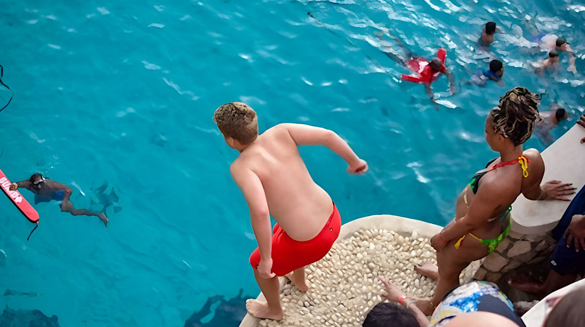 Boy in red swim trunks diving from a pebble-topped ledge into a bright turquoise outdoor pool while friends in swimsuits watch and swimmers paddle below.