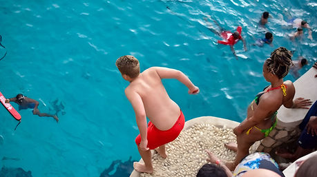 Boy in red swim trunks diving from a pebble-topped ledge into a bright turquoise outdoor pool while friends in swimsuits watch and swimmers paddle below.