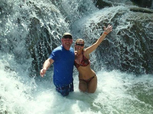 Two people standing waist-deep in a frothy pool beneath a cascading waterfall, smiling and waving — man in a blue shirt and cap and woman in a patterned bikini with sunglasses.