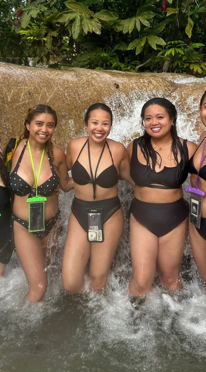 Five women in swimsuits posing and smiling under a small tropical waterfall in a natural pool, wearing waterproof phone pouches with lush green jungle foliage behind them.