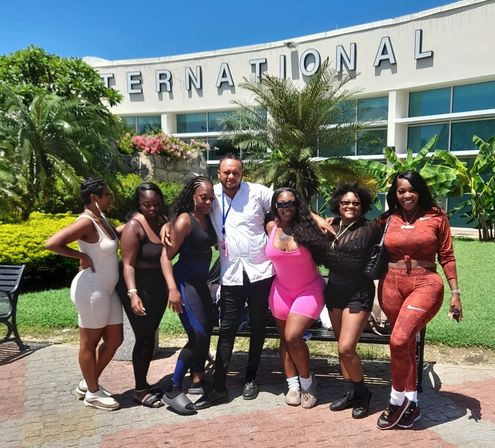 Group of seven travelers posing outside an international terminal on a sunny day with palm trees and tropical landscaping, wearing colorful casual outfits for a vacation.