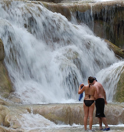 Couple in swimwear standing at the base of a multi-tiered waterfall, water rushing over limestone rock terraces into a shallow pool