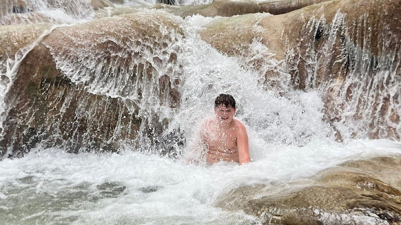 Person sitting under a cascading natural waterfall on smooth rocky terraces, laughing in a frothy pool of water