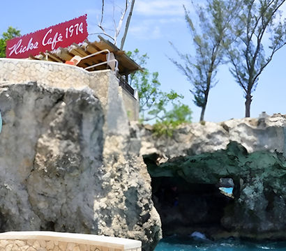 Cliffside seaside café scene with a colorful round sign, rugged limestone cliffs and a rocky sea cave opening to turquoise water beneath a bright blue sky and windswept tropical trees.