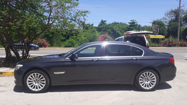 Dark gray luxury sedan parked curbside on a sunny road, framed by trees and pink flowering shrubs with an open SUV trunk in the background.