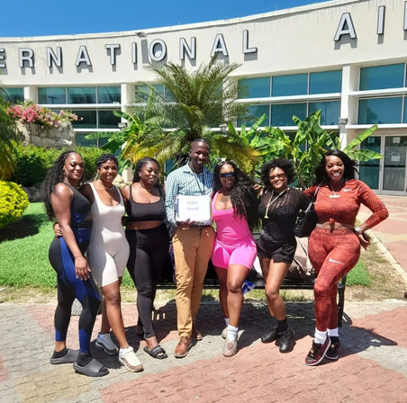 Group of six women and one man smiling and posing outside an international airport entrance with palm trees and tropical landscaping on a sunny day.