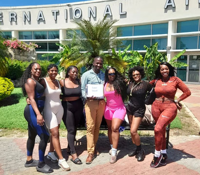 Group of six women and one man smiling and posing outside an international airport entrance with palm trees and tropical landscaping on a sunny day.