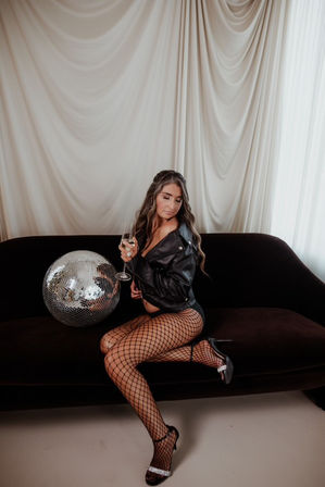 Edgy maternity photoshoot: pregnant model in a black leather jacket, fishnet tights and heels sits on a dark sofa with a wine glass beside a disco ball against draped white curtains.