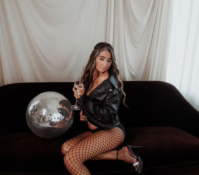 Edgy maternity photoshoot: pregnant model in a black leather jacket, fishnet tights and heels sits on a dark sofa with a wine glass beside a disco ball against draped white curtains.