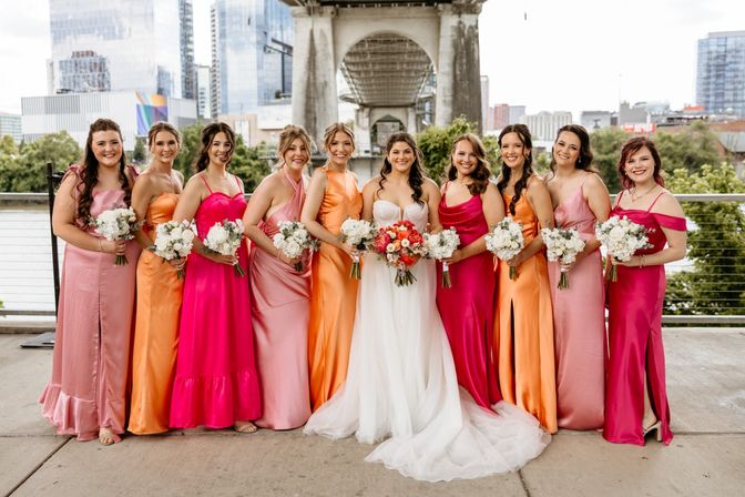 Bride in a white gown with ten bridesmaids in pink, fuchsia, and orange satin dresses holding bouquets, posing under an urban bridge with a river and city skyline in the background.