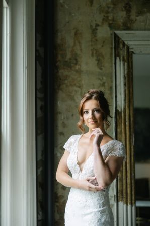 Elegant bride in a lace wedding gown standing by a tall window with soft natural light, hand gently touching her chin, vintage textured wall and mirror in the background — intimate bridal portrait.