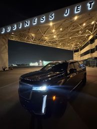 Black SUV parked under an illuminated "BUSINESS JET" terminal canopy at a private jet hangar on an airport ramp at night, with hangar lights and distant aircraft visible