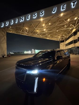 Black SUV parked under an illuminated "BUSINESS JET" terminal canopy at a private jet hangar on an airport ramp at night, with hangar lights and distant aircraft visible
