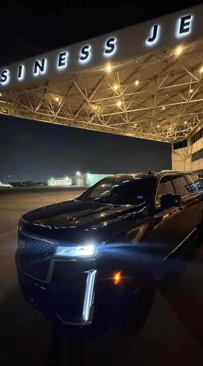 Black SUV parked under an illuminated "BUSINESS JET" terminal canopy at a private jet hangar on an airport ramp at night, with hangar lights and distant aircraft visible