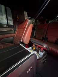 Cozy SUV interior with brown leather seats and embroidered headrests, center console opened to reveal an ice-filled cooler with champagne and liquor bottles, suburban houses visible through the window.