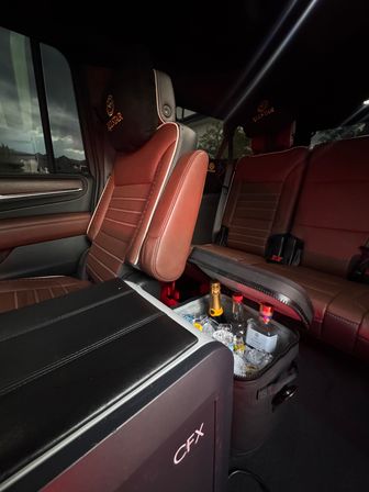Cozy SUV interior with brown leather seats and embroidered headrests, center console opened to reveal an ice-filled cooler with champagne and liquor bottles, suburban houses visible through the window.