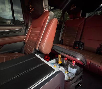 Cozy SUV interior with brown leather seats and embroidered headrests, center console opened to reveal an ice-filled cooler with champagne and liquor bottles, suburban houses visible through the window.