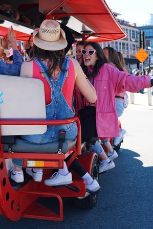 Group of friends laughing on a red pedal-pub in a sunny downtown street, woman in denim overalls and straw cowboy hat, others wearing pink jacket and heart-shaped sunglasses.