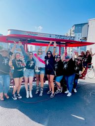 Smiling group of women cheering on a red Nashville party bike during a bachelorette outing, wearing casual summer outfits and cowboy boots with city skyline in the background.