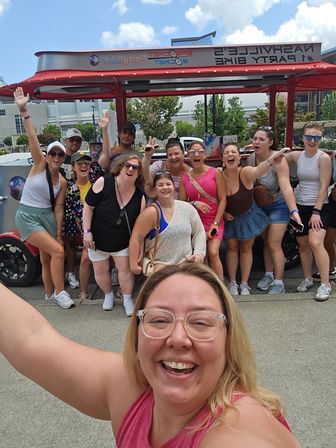 Large group of friends taking a cheerful group selfie in front of a red Nashville party bike on a sunny day