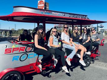 Group of women laughing and holding drinks on a red pedal-powered party bike at the Nashville riverfront, sunny blue sky with a bridge and stadium in the background.