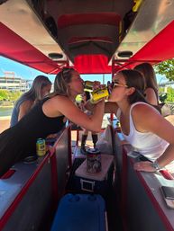 Two women clinking canned drinks on a red-canopied pedal pub along an urban riverside, friends enjoying a sunny outdoor group outing near a bridge.