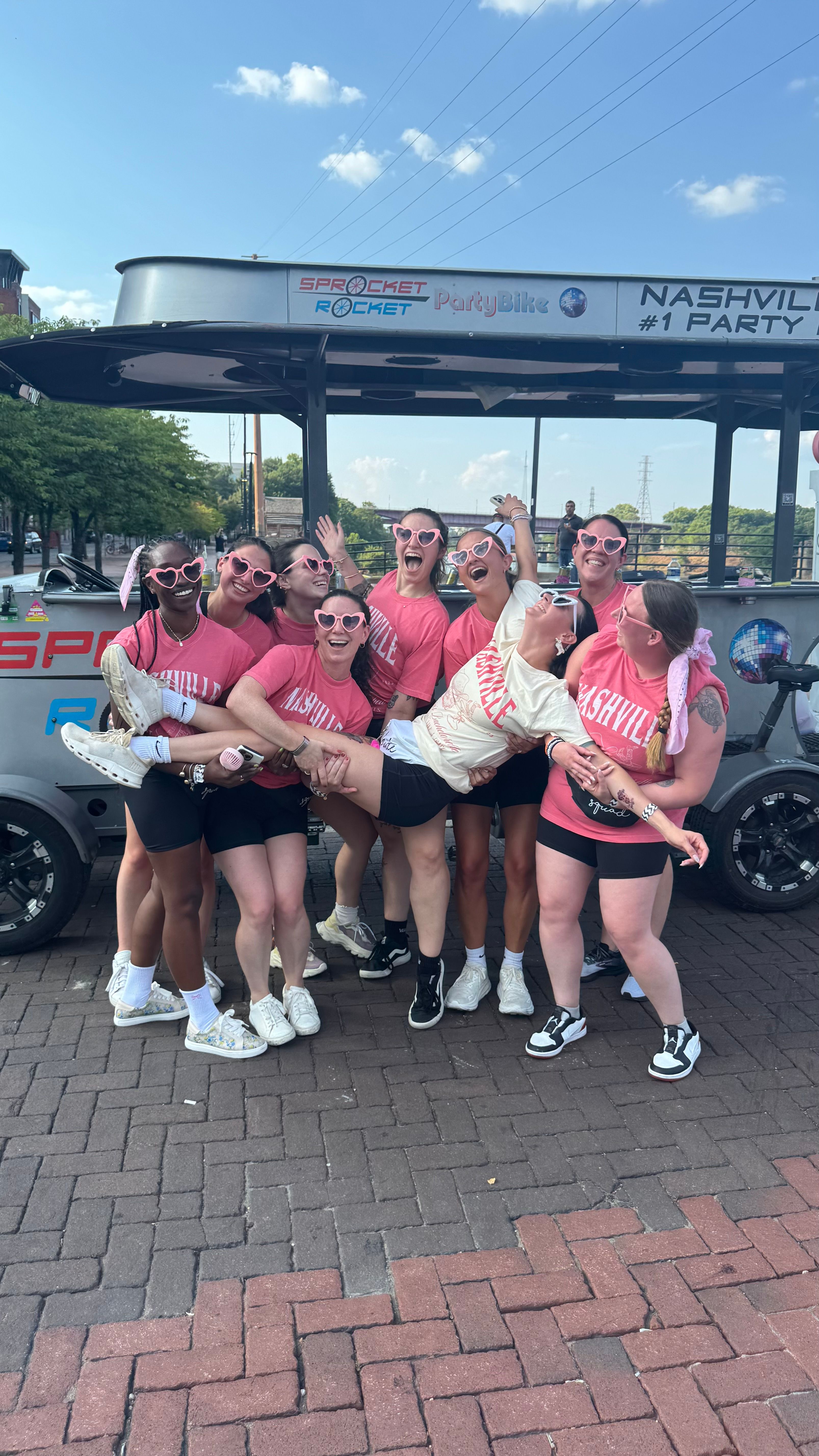 Energetic group of friends in matching pink 'Nashville' shirts and heart-shaped sunglasses lifting a friend in front of a party bike on a sunny downtown brick street.