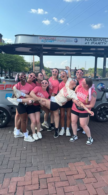 Energetic group of friends in matching pink 'Nashville' shirts and heart-shaped sunglasses lifting a friend in front of a party bike on a sunny downtown brick street.