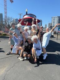 A joyful group wearing white curly wigs and vintage-style floral dresses cheering and posing under a red canopy in a sunny urban parking lot with city skyline in the background.