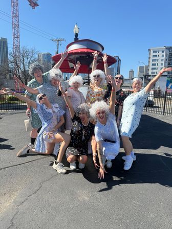 A joyful group wearing white curly wigs and vintage-style floral dresses cheering and posing under a red canopy in a sunny urban parking lot with city skyline in the background.