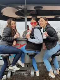 Three friends on a riverside pedal bar seated on bike-style stools, sipping canned drinks through curly straws — one wearing pink heart sunglasses, casual jackets and jeans near a bridge.