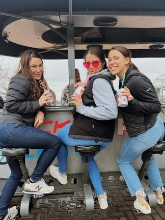 Three friends on a riverside pedal bar seated on bike-style stools, sipping canned drinks through curly straws — one wearing pink heart sunglasses, casual jackets and jeans near a bridge.