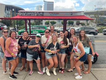 Smiling group of friends celebrating on a downtown Nashville sidewalk, posing in front of a red pedal-powered party bike while wearing casual summer outfits and holding drinks on a sunny day.