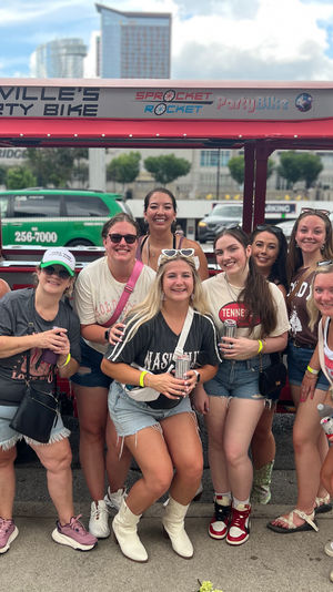Smiling group of friends celebrating on a downtown Nashville sidewalk, posing in front of a red pedal-powered party bike while wearing casual summer outfits and holding drinks on a sunny day.