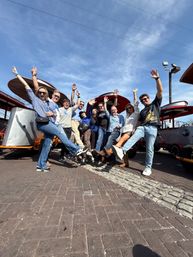 Group of smiling adults in casual clothes kicking legs and raising hands beside red-roofed pedal-party vehicles on a sunny downtown cobblestone street under a bright blue sky