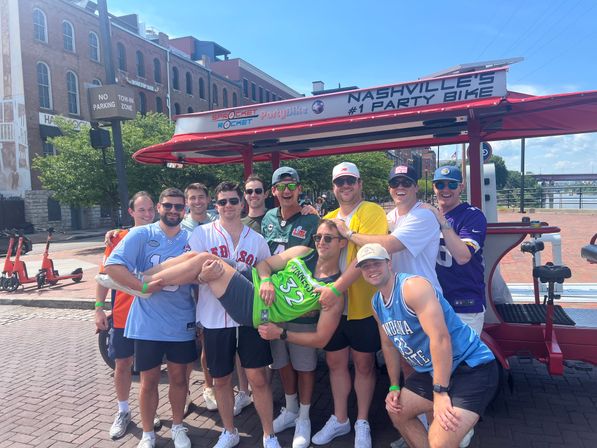 Ten friends in sports jerseys and sunglasses smiling and holding a teammate in front of a red party bike on a sunny day by the downtown Nashville riverfront brick street.
