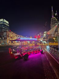 Neon-lit party bike parked on a brick sidewalk in downtown Nashville at night, purple and red lights glowing with the city skyline and tall spired tower in the background.