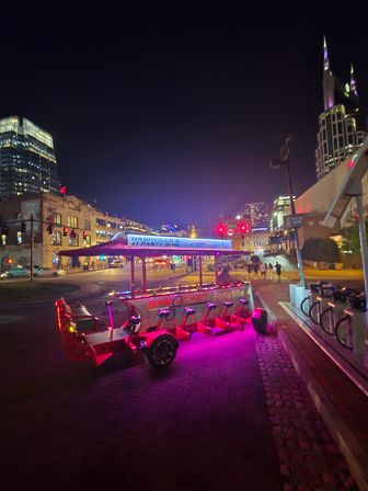 Neon-lit party bike parked on a brick sidewalk in downtown Nashville at night, purple and red lights glowing with the city skyline and tall spired tower in the background.