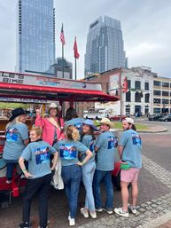 Smiling group in matching blue T‑shirts and cowboy hats riding a red pedal party bike through a downtown streetscape with glass skyscrapers, brick bars and American flags